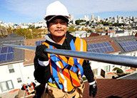 Construction worker wearing hard hard and holding metal pipe with roofs containing solar panels in the background.